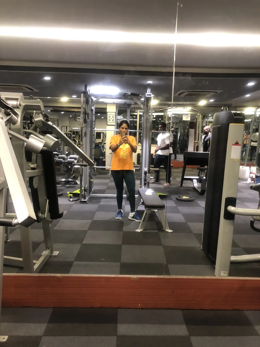 Gym floor with machines and a mirror, a person in an orange top standing in the middle