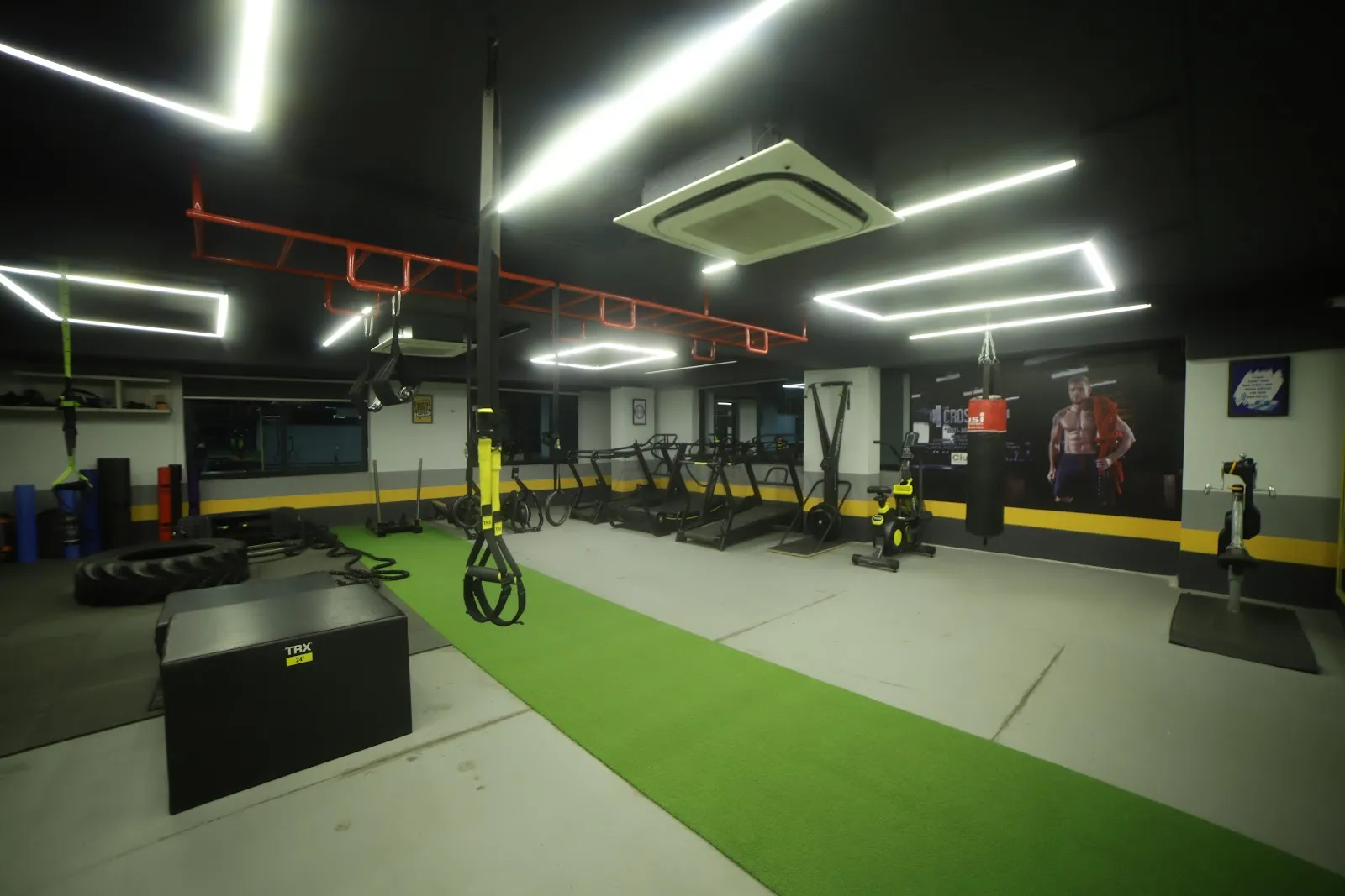 Training floor with a green turf strip, a black plyo box, rigs and a boxing bag under ceiling light strips
